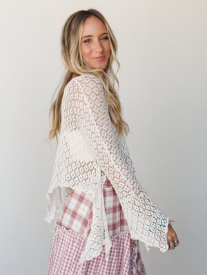 A model glances back over her shoulder in a cream open-weave crochet poncho layered over a pink gingham tiered skirt — the kind of effortless mix that looks thrown together in the best possible way.