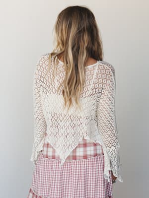 Shot from behind, a model wears a cream open-knit crochet poncho over a pink gingham skirt — the kind of effortless layering that looks like it just happened.