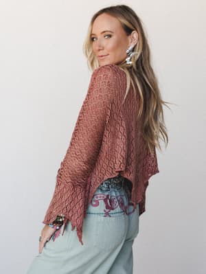 A model glances over her shoulder in a dusty rose open-knit crochet poncho layered over embroidered sage jeans — effortless boho layering with a lived-in, free-spirited feel.
