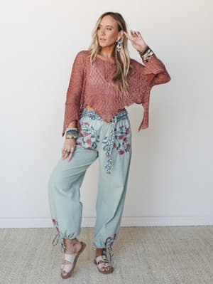 A relaxed boho look built around a dusty rose open-knit crochet poncho top paired with sage green floral-print harem pants, styled with stacked bracelets and flat sandals against a soft neutral studio wall.