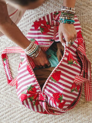 An overhead lifestyle shot of a quilted strawberry-print duffle bag being unzipped by hands stacked with colorful beaded bracelets and rings — the kind of bag that makes packing feel like the best part of the trip.