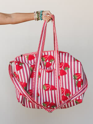 A hand holds up an oversized quilted tote bag in a cheerful strawberry-and-stripe print — the kind of bag that makes a farmers market run feel like a whole personality.