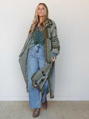 A long, embroidered olive duster coat styled open over a knotted sage henley and wide-leg light wash jeans, grounded with leopard print ankle boots — the whole look has that effortless, collected energy of someone who got dressed without overthinking