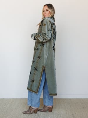A floor-length embroidered velvet coat in dusty sage layered over straight-leg jeans and leopard print booties — the kind of statement outer layer that makes a whole outfit feel intentional without trying.