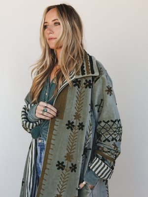 A long embroidered olive jacket layered over a sage henley and jeans, styled with silver and turquoise rings — the kind of piece that looks like it has a whole history and smells like cedar.