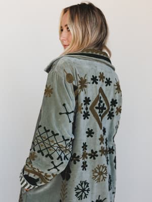 A model shot from behind showing a dusty sage velvet jacket covered in intricate black and gold embroidery — floral motifs, diamond shapes, and folk-inspired symbols that feel like a vintage find with serious intention behind it.