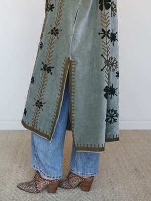 A close-cropped lower-body shot showing a heavily embroidered sage velvet duster layered over light wash straight jeans and leopard print ankle boots — the kind of outfit that makes a Tuesday feel intentional.