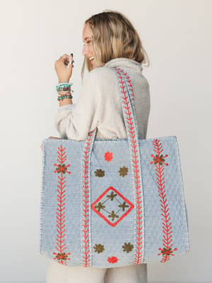 A laughing model shown from behind carries an oversized quilted tote in dusty blue, covered in coral embroidered vines and olive floral motifs — the kind of bag that makes errands feel like a little adventure.