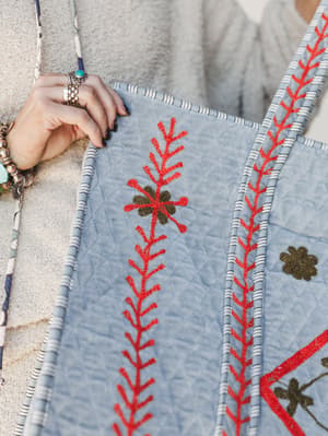 A close-up shot of a quilted denim blue duffle bag with bold red embroidered botanical stitching and olive floral appliqué, held by a model layered in silver and turquoise rings and stacked bracelets — the kind of bag that looks like it has a story b