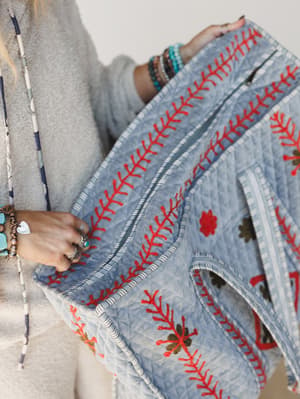 A close-up of a quilted denim blue duffle bag held by a model layered in stacked bracelets and rings — the red embroidered branch motifs and floral appliqués give it that well-traveled, picked-it-up-somewhere-special energy.
