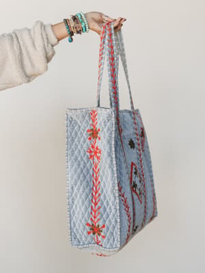 A quilted denim blue tote bag with coral red embroidery and floral motifs held up by a hand layered in boho beaded bracelets — the kind of bag that looks like it belongs at a farmers market or a weekend road trip.
