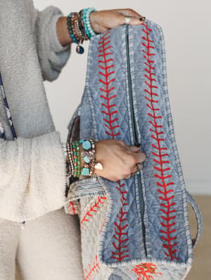 A close-up of hands holding a quilted denim blue duffle bag with bold red embroidery stitching, styled against a cozy cream sweater and stacked turquoise and mixed bead bracelets — the kind of bag that makes even a parking lot feel like a destination