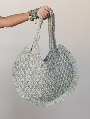 A round quilted tote in sage green floral block print with a striped ruffle trim, held up by a beaded-bracelet-stacked wrist — the kind of bag that makes a farmers market run feel like a whole thing.