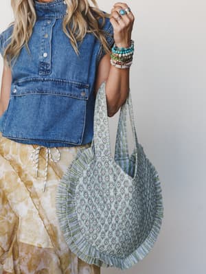 A close-up lifestyle shot of a model holding a large round printed tote bag in sage and cream, styled with a sleeveless denim top, a flowy tie-dye skirt, and a stacked arm of beaded and stone bracelets — the whole look feels like a very good Saturday