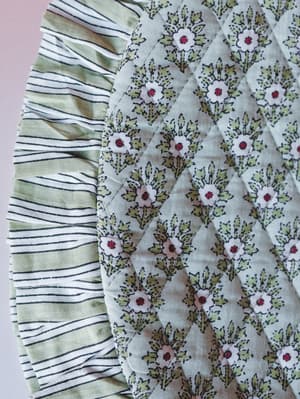 A close-up of layered fabric panels showing two distinct prints side by side — a sage and cream stripe alongside a quilted floral bouquet print in soft green, white, and cranberry — shot against a blush background with a fresh, garden-y feel.
