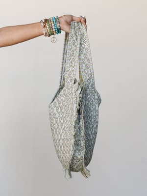 A hand adorned with stacked beaded bracelets and a peace charm holds up a roomy printed tote bag — the kind that looks equally at home at the farmers market or the beach.