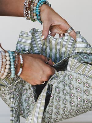 A close-up of two beaded-bracelet-stacked hands holding open a quilted floral and stripe tote bag in soft sage and cream — the kind of bag that looks like it already has a farmers market receipt and a lip balm inside it.