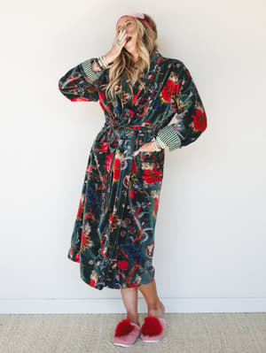 A laughing blonde model wears a floor-length velvet floral robe in deep forest green with bold botanical print, striped cuff contrast, and a tie waist — styled with a pink sleep mask headband and fluffy red slippers for a look that makes staying home