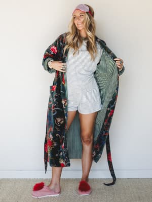A smiling model holds open a dramatic floor-length floral velvet robe in deep green, layered over a grey striped romper with fuzzy pink slippers and a gingham sleep mask — the whole look has the energy of a very stylish Sunday morning.