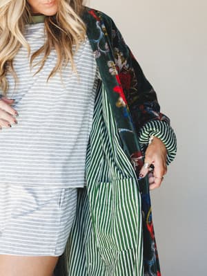 A close-cropped shot of a woman layering a dramatic floral velvet-trimmed duster jacket with a green and white striped lining over a heathered grey striped sweatshirt set — the mix of prints feels intentional and a little unexpected, in the best way.