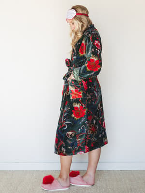 A velvet floral robe in deep forest green with bold red blooms is styled with a satin sleep mask and fluffy pink slippers — the kind of morning-at-home look that feels intentional rather than accidental.