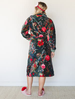 Shot from the back, a model wears a lush velvet floral robe in deep forest green with bold botanical print, styled with a red ruffle headband and fuzzy pink slippers — the kind of cozy that doesn't apologize for itself.