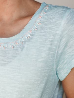 A close-up of a soft aqua slub-knit tee with a sweet detail worth leaning in for — hand-stitched whipstitch embroidery in blush pink and cream tracing the neckline.
