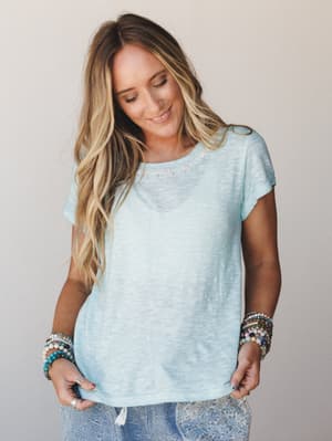 A relaxed aqua slub-knit tee with delicate neckline embroidery, styled with stacked beaded bracelets and lace-print pants — the kind of effortless summer look that feels good without trying too hard.