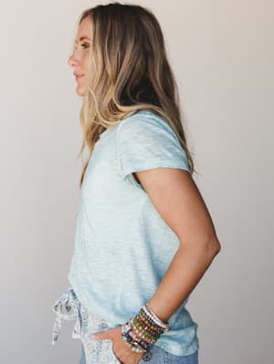 A side-profile shot of a model in a soft aqua slub-knit tee, styled with lace-trim bottoms and a stacked beaded bracelet set — the kind of easy, collected look that feels just as good as it looks.