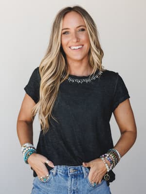 A sun-kissed model in a washed black crew-neck tee with subtle neckline embellishment, layered with stacks of beaded and charm bracelets on both wrists — effortless and a little collected, like she got dressed without trying too hard.