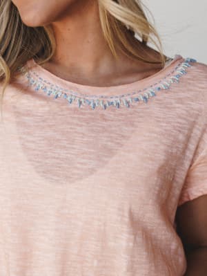 A close-up detail shot of a dusty peach slub-knit tee with delicate blue and mint embroidery stitching along the neckline — the kind of top that looks effortless but clearly has something special going on.