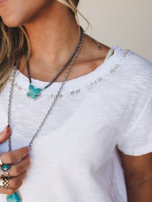 A close-up lifestyle shot of a white slub-knit tee with delicate embroidery at the neckline, styled with layered silver and turquoise jewelry — effortless and a little sentimental, like a favorite shirt you've had forever.