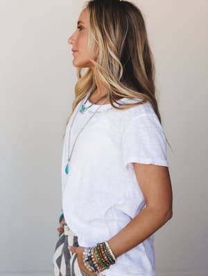 A side-profile shot of a model in a relaxed white slub tee, layered with turquoise stone necklaces and a stacked beaded bracelet set — the kind of effortless that takes exactly the right accessories.