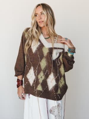 An oversized argyle sweater in warm mocha tones layered over a white lace skirt, styled with stacked turquoise and leather bracelets and layered necklaces — the kind of outfit that looks like it came together effortlessly but absolutely did not.