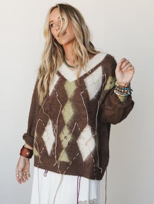 An oversized argyle knit sweater in mocha and sage layered over a white lace midi skirt, styled with stacked boho jewelry — the kind of outfit that looks like it came together effortlessly but absolutely did not.