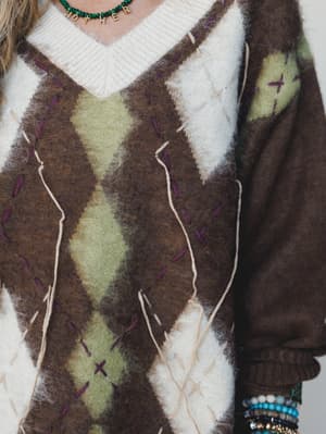 A close-up detail shot of a fuzzy argyle sweater in mocha, sage, and cream — the kind of piece that makes you want to curl up somewhere with a warm drink and good company, styled with a beaded letter necklace and stacked bracelets.
