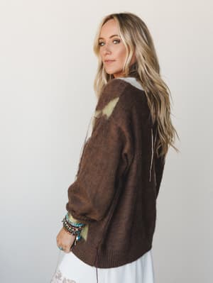 Shot from a three-quarter back angle, a model looks over her shoulder in a cozy oversized mocha sweater with sage green paint-stroke detailing, styled with stacked boho bracelets and a white skirt peeking out below — the whole thing feels like autumn