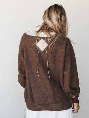 Shot from behind, a model in a chunky mocha knit sweater shows off the standout back detail — a cream diamond patch with dangling string ties — layered over a white skirt with a leather cuff bracelet adding just the right amount of character.