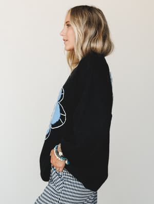 A side-profile shot of a model in a cozy black sweater with oversized blue floral intarsia, paired with gingham pants and a stack of eclectic bracelets — the kind of outfit that looks effortless but definitely wasn't an accident.