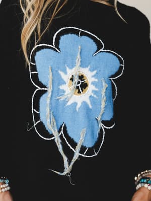 A close-up detail shot of a black sweater featuring an oversized appliqué flower in periwinkle blue with raw-edge fringe stems and white embroidered outline — the kind of sweater that looks like wearable folk art and feels like a personality statemen