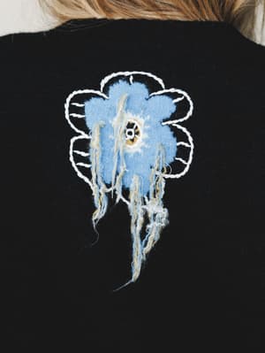 A close-up of the back of a black sweater featuring a handcrafted 3D flower appliqué in cornflower blue with white chain-stitch petal outlines and trailing fringe threads — the kind of detail you notice across the room.