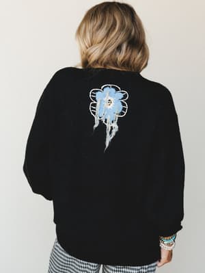 A back-facing shot of a cozy black sweater with a standout frayed floral appliqué detail — blue flower with raw, trailing threads — styled with gingham pants and a stack of mixed bracelets for that effortlessly personal, lived-in feel.