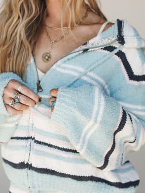A close-up of a soft, striped knit hoodie in sky blue, cream, blush, and navy — held open at the zip by a hand stacked with silver and turquoise rings, layered gold necklaces peeking out at the neckline, the whole thing feeling like a lazy morning wi