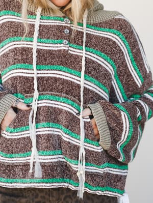 A close-up shot of a chunky striped knit hoodie in warm chocolate brown with kelly green and cream stripe detailing — the kind of sweater that looks like it was borrowed from someone with great taste.