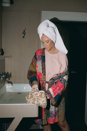A woman with a towel-wrapped hair stands at a vintage farmhouse sink, layering a bold patchwork kimono jacket over a dusty pink waffle top — the whole scene feels like a very stylish morning routine.
