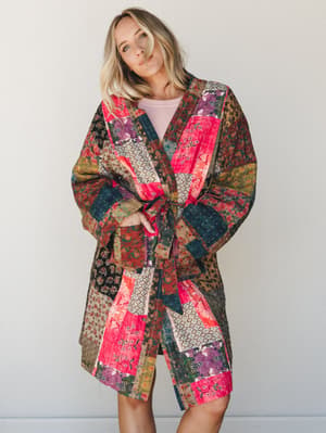 A quilted patchwork kimono jacket in a riot of floral and geometric prints — hot pink, teal, and earth tones — belted loosely over a soft pink top, with a relaxed, one-of-a-kind energy that feels like it came from a very well-traveled vintage shop.