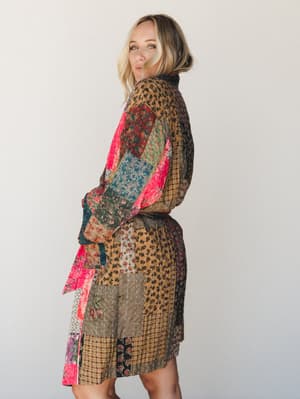 A blonde model poses from a three-quarter back angle in a patchwork kantha-style jacket — earthy tans and olives layered with pops of hot pink, the whole thing stitched together like a quilt someone actually wants to wear.