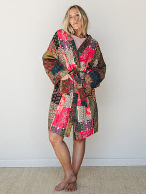 A patchwork quilted kimono-style jacket in a bold mix of floral and geometric patches — hot pink, teal, and earthy tones — worn open with a tie belt over a blush pink top, styled barefoot against a soft neutral wall for an effortless, lived-in boho f