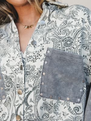 A close-up of a grey paisley button-snap jacket with mixed fabric detailing — the intricate floral and scroll print contrasts against a waffle-knit chest pocket, styled with a delicate gold necklace for that effortless, put-together feel.