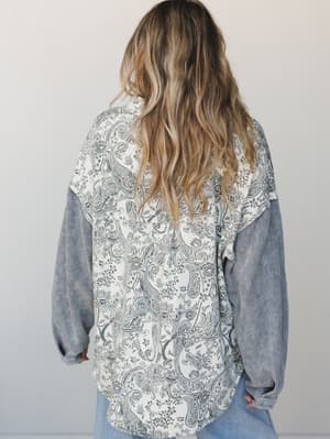 Shot from behind, a model wears a relaxed mixed-fabric jacket with a bold paisley-printed back panel and contrasting grey waffle-knit sleeves, paired with light wash jeans — the kind of piece that does the talking for you.
